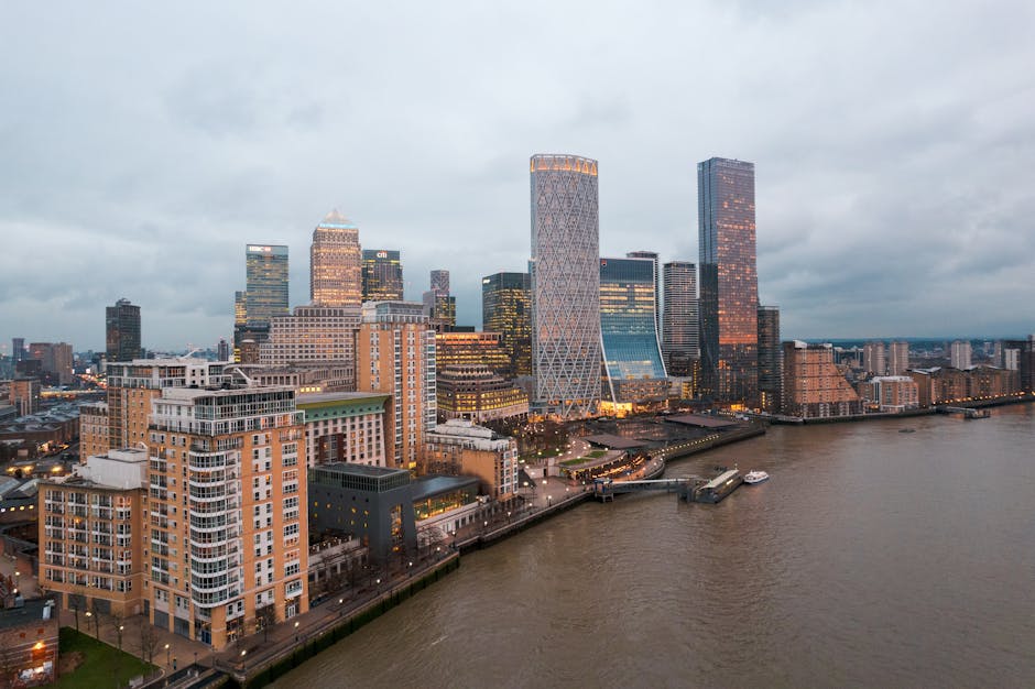 An aerial view of a modern city skyline along a wide river, featuring a mix of tall skyscrapers with glass facades and some with brick or stone exteriors. Prominent buildings include a distinctive tow