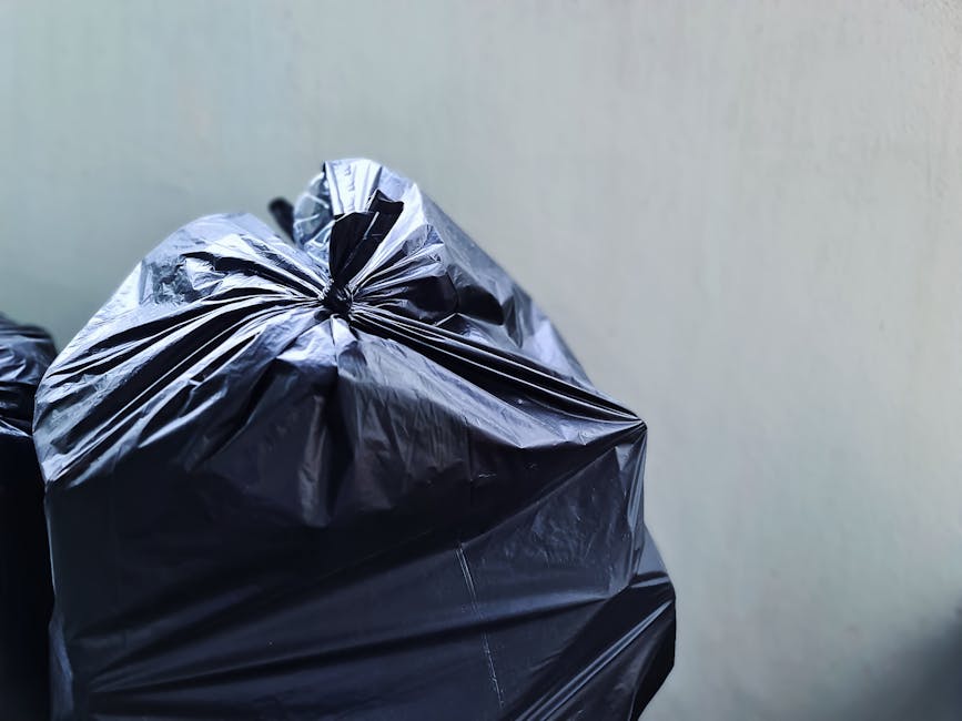 A collection of overflowing waste and rubbish bags piled on a paved city street corner near a metal railing, including black plastic refuse bags, cardboard boxes, paper and packaging waste, and loose 